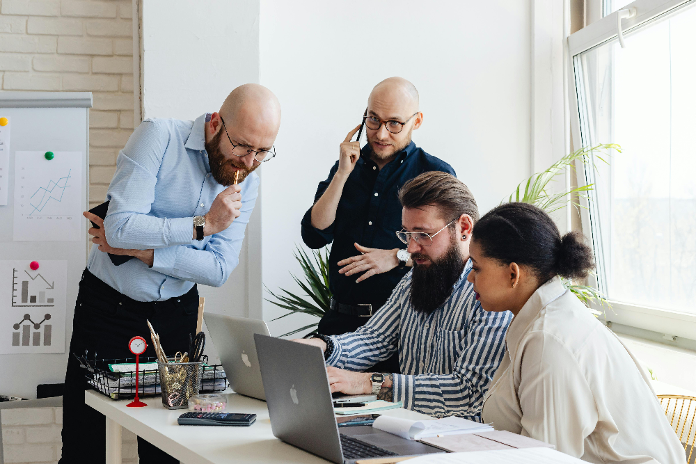 A group of four professionals collaborating around a desk with laptops and charts in a sunlit, modern office space.